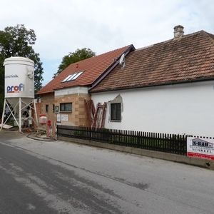 Haus mit rotem Ziegeldach und teilweise renovierter Fassade an einer Straße. Links steht ein großer Bausilo, rechts ist ein Bauzaun und ein Renovierungsschild sichtbar.