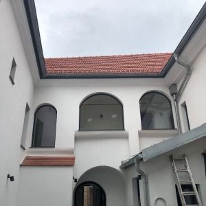 Innenhof eines frisch renovierten Gebäudes mit weißer Fassade, roten Dachziegeln und rundbogigen Fenstern. Eine Leiter lehnt an der Wand im unteren Bereich.