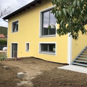 Gelbes Wohnhaus mit großem Fenster und Außentreppe, Neubau in Hanglage.