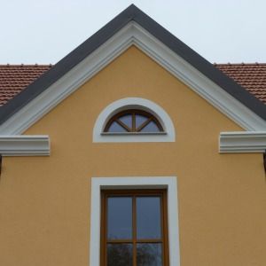 Giebelansicht eines Hauses mit ockerfarbener Fassade, halbkreisförmigem Fenster und grauem Ziergiebel