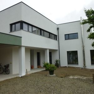 Großzügiger, weißer Neubau mit mehreren Fensterflächen, Flachdach und einem begrünten Hof mit Kiesboden.