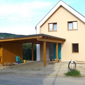 Einfamilienhaus mit gelber Putzfassade, spitzem Satteldach und überdachter Holzterrasse. Ein Fahrrad steht neben dem Eingang.