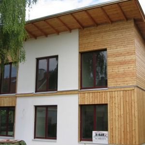 Zweigeschossiges Wohnhaus mit weißer Fassade, dunklen Fensterrahmen und Holzverkleidung im oberen Bereich, unter einem Flachdach mit Überstand.