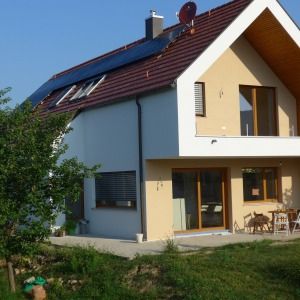 Einfamilienhaus mit rotem Satteldach, weißer Fassade und überdachter Terrasse. Der Garten im Vordergrund ist grün bepflanzt.