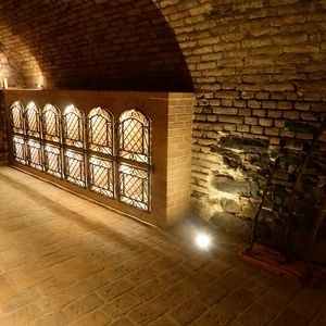Gewölbegang mit Backsteinmauer, beleuchtet durch Bodenstrahler vor verzierten Fensteröffnungen