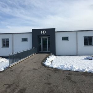Flacher Gewerbebau mit grauer und weißer Fassade, leicht ansteigender Zugang und Schnee im Vordergrund