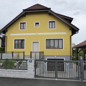 Zweigeschossiges Haus mit gelber Fassade, dunkelbraunem Dach und grauem Zaun vor dem Grundstück