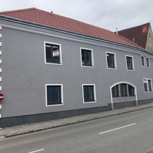 Wohnhaus mit grauer Fassade, rotem Ziegeldach und mehreren Fenstern, direkt an einer Straße gelegen