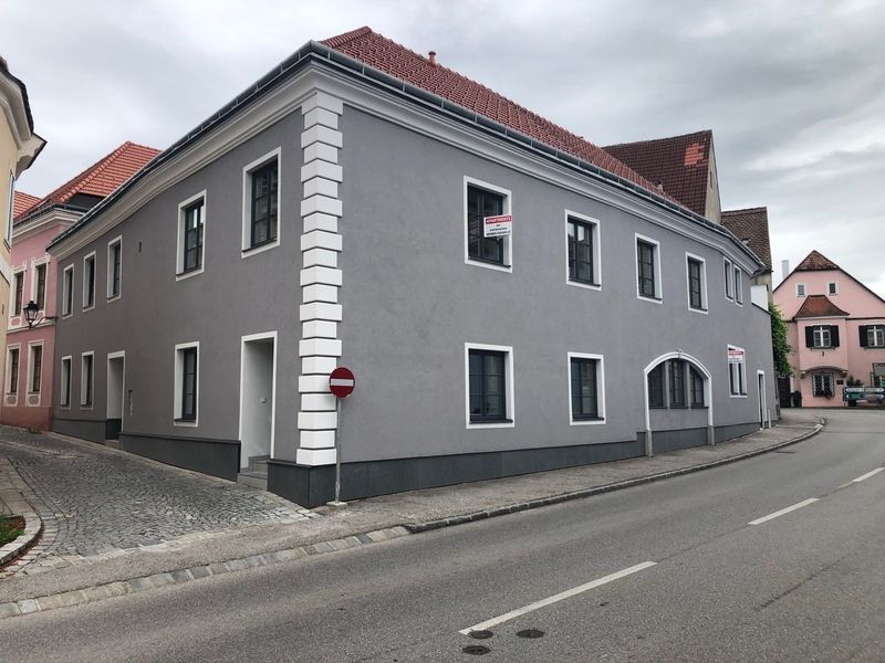 Zweigeschossiges Eckhaus mit grauer Fassade, weißen Fensterrahmen und roten Dachziegeln. Die Hausecken sind mit weißen Zierleisten betont.