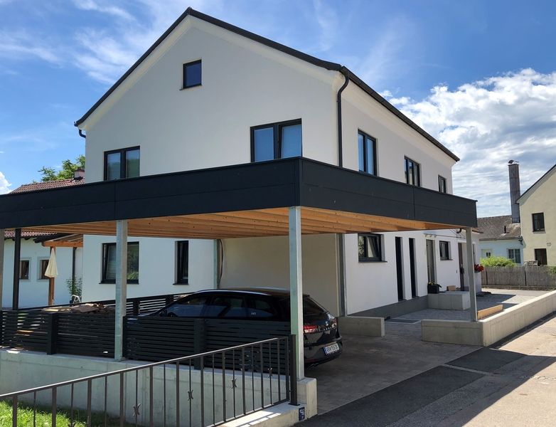 Zweigeschossiges Wohnhaus mit weißer Fassade, schwarzem Flachdach und integriertem Carport mit Holzdecke. Ein schwarzes Auto steht unter dem Carport, davor ein niedriger Betonsockel mit Zaun. Die Fensterrahmen und Türen sind dunkel gehalten, moderne Bauweise.