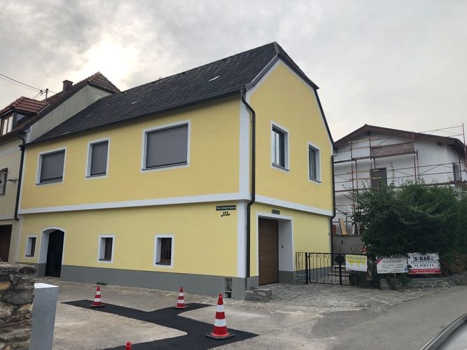 Zweigeschossiges Haus mit hellgelber Fassade, weiß abgesetzten Fensterrahmen und dunklem Dach. Der Eingangsbereich ist über wenige Stufen erreichbar, vor dem Haus stehen mehrere Baustellenkegel und im Hintergrund ein eingerüstetes Nachbarhaus.
