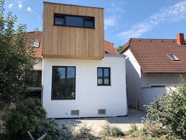 Haus mit weißer Putzfassade und rotem Ziegeldach, modern erweitert durch einen würfelförmigen Holzaufbau im oberen Bereich. Große Fenster an der Vorderseite, umgeben von Büschen und Pflanzen im Garten.