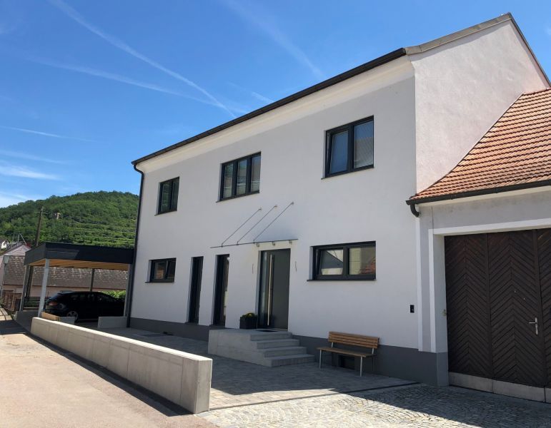 Modernisiertes Wohnhaus mit weißer Fassade und rotem Ziegeldach. Schwarze Fensterrahmen, eine überdachte Eingangstür mit Stufen und eine kleine Holzbank vor dem Gebäude. Links daneben befindet sich ein Carport.